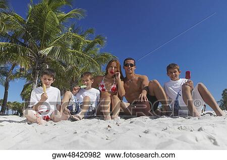 Stock Image - Mid adult man and a young man sitting with their children on the beach. Fotosearch