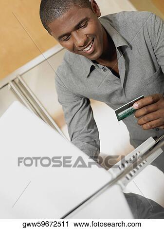 Stock Image - Mid adult man holding a credit card in front of a laptop. Fotosearch