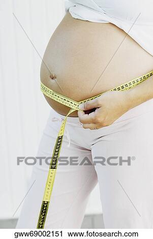 Stock Image - Mid section view of a pregnant woman measuring her abdomen with a tape measure. Fotosearch