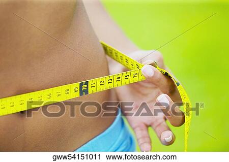 Stock Image - Mid section view of a woman measuring her waist with a tape measure. Fotosearch