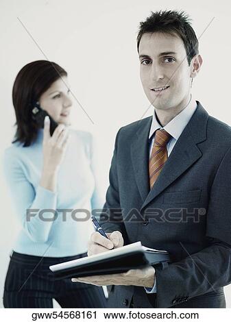 Portrait of a businessman holding a personal organizer with a businesswoman talking on a mobile phone in the background View Large Photo Image Stock Image - Portrait of a businessman holding a personal organizer with a businesswoman talking on a mobile phone in the background. Fotosearch