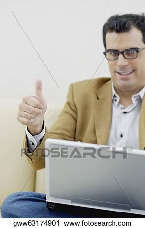 Stock Image - Portrait of a mid adult man sitting with a laptop and giving a thumbs up sign. Fotosearch