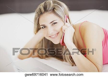 Stock Image - Portrait of a mid adult woman lying in front of a laptop and talking on a mobile phone. Fotosearch