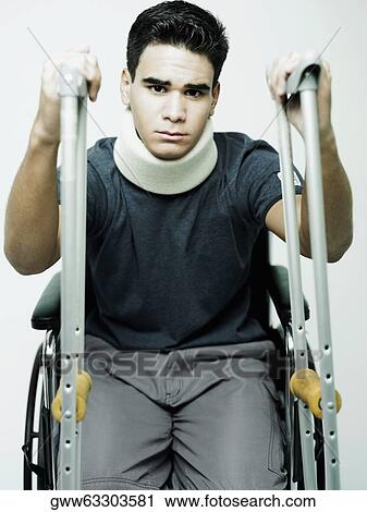 Stock Image - Portrait of a young man sitting in a wheelchair and holding a pair of crutches. Fotosearch