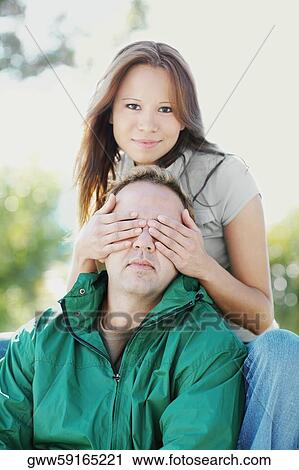 Stock Image - Portrait of a young woman covering a mid adult man's eyes. Fotosearch