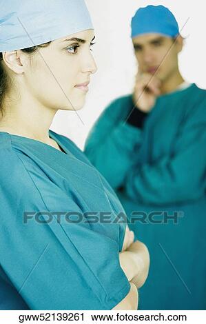 Stock Image - Side profile of a female doctor standing with her arms crossed and a male doctor standing in the background. Fotosearch