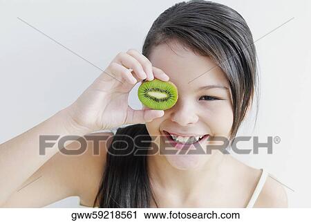 Stock Image - Teenage girl holding a slice of kiwi fruit in front of her eye. Fotosearch