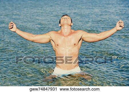 Stock Image - Young man standing in water with his arms outstretched. Fotosearch