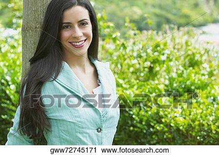 Young woman leaning against a tree trunk in a park and smiling View Large Photo Image Stock Image - Young woman leaning against a tree trunk in a park and smiling. Fotosearch