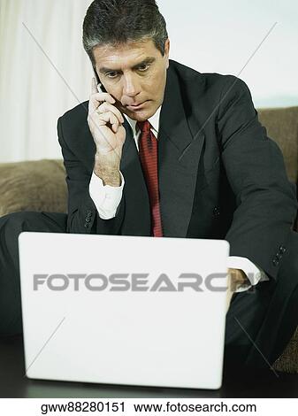 Stock Image - Close-up of a businessman talking on a mobile phone and operating a laptop. Fotosearch