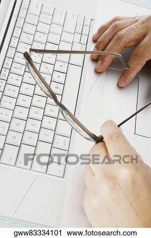 Stock Image - Close-up of a person's hand holding eyeglasses and using a laptop. Fotosearch