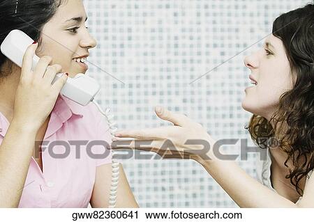 Close-up of a young woman using a telephone with her friend sitting in front of her View Large Photo Image Stock Image - Close-up of a young woman using a telephone with her friend sitting in front of her. Fotosearch