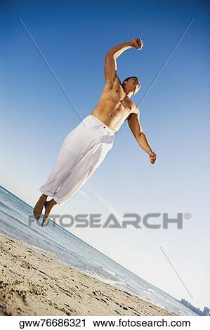Stock Image - Low angle view of a young man jumping on the beach with his hand raised . Fotosearch