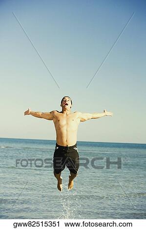 Stock Image - Mid adult man jumping on the beach with his arms outstretched . Fotosearch