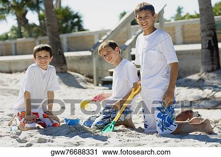 Stock Image - Portrait of two boys and a teenage boy playing with sand pail and shovel on the beach. Fotosearch