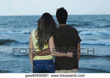 Stock Image - Rear view of a young couple standing on the beach. Fotosearch