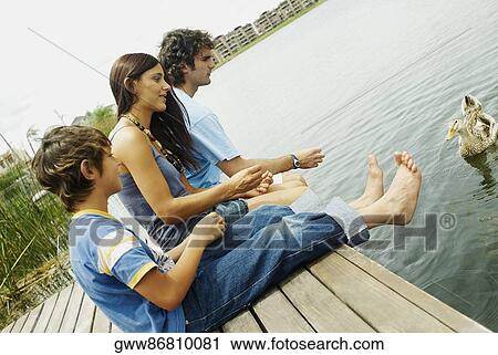 Stock Image - Side profile of a boy with his parents feeding a duck at a lake. Fotosearch