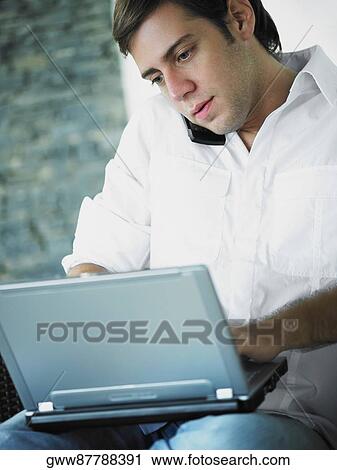 Stock Image - Young man using a laptop while talking on a mobile phone. Fotosearch