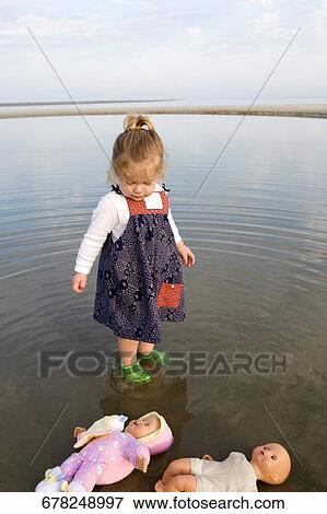 Stock Photo - Baby girl standing in water and looking at her dolls. Fotosearch