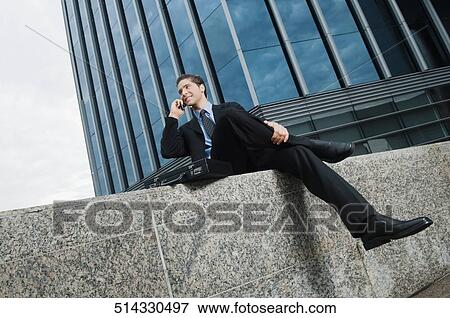 Businessman sitting at the edge of a wall and talking on a mobile phone View Large Photo Image Stock Photo - Businessman sitting at the edge of a wall and talking on a mobile phone. Fotosearch
