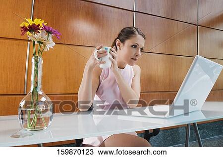 Stock Image - Businesswoman holding a coffee cup and looking at a laptop. Fotosearch