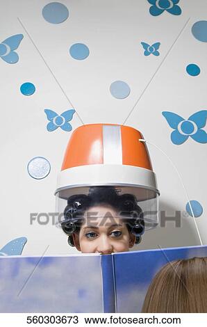 Close-up of a young woman sitting under hair dryer and reading a magazine View Large Photo Image Stock Image - Close-up of a young woman sitting under hair dryer and reading a magazine. Fotosearch