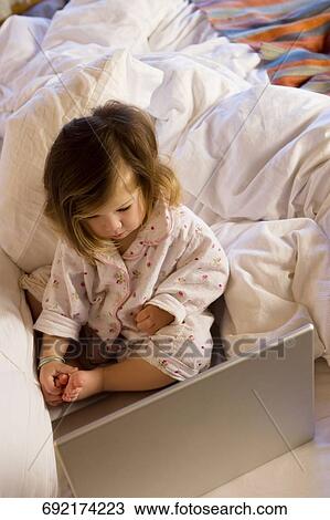 Stock Image - High angle view of a girl sitting on the bed and looking at a laptop. Fotosearch