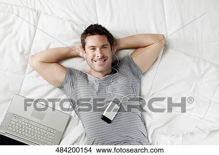 Picture - High angle view of a mid adult man lying on the bed and listening to music. Fotosearch