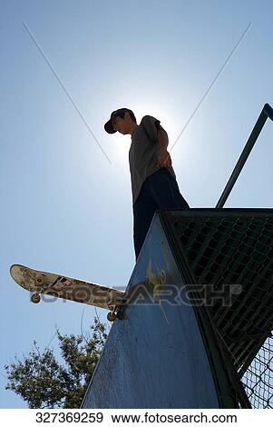 Low angle view of a young male skateboarder standing on a stunt ramp View Large Photo Image Stock Photo - Low angle view of a young male skateboarder standing on a stunt ramp. Fotosearch