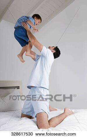 Stock Image - Mid adult man playing with his son on the bed. Fotosearch