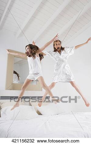 Stock Image - Mid adult woman jumping with her daughter on the bed. Fotosearch