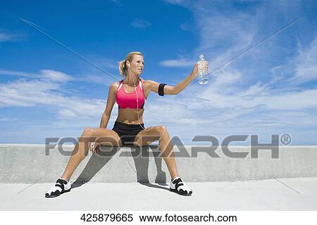 Stock Photography - Mid adult woman sitting on a ledge and holding a water bottle. Fotosearch