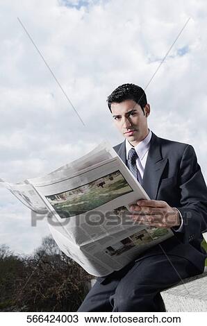 Stock Image - Portrait of a businessman sitting at the edge of a wall and reading a newspaper. Fotosearch