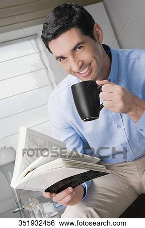 Stock Photograph - Portrait of a mid adult man holding a book and drinking coffee. Fotosearch