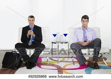 Stock Photo - Portrait of a young man and a mid adult man sitting on chairs and holding mobile phones. Fotosearch
