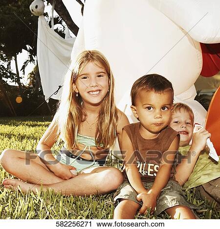 Stock Image - Portrait of three children sitting in the lawn and smiling. Fotosearch