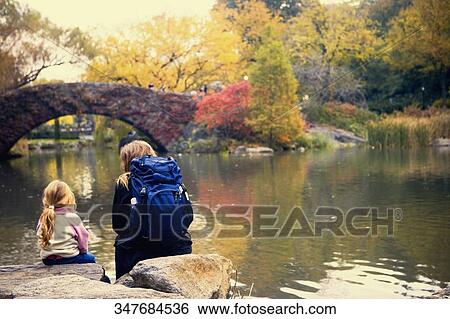 Rear view of a girl and a woman sitting on rocks along a river, New York City, New York State, USA View Large Photo Image Stock Photograph - Rear view of a girl and a woman sitting on rocks along a river, New York City, New York State, USA. Fotosearch