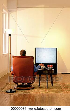 Rear view of a man sitting in an armchair and watching television View Large Photo Image Stock Photograph - Rear view of a man sitting in an armchair and watching television. Fotosearch