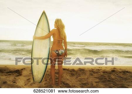 Stock Image - Rear view of a young woman with a surfboard standing on the beach. Fotosearch