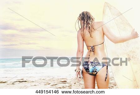 Stock Photograph - Rear view of a young woman with a surfboard standing on the beach. Fotosearch
