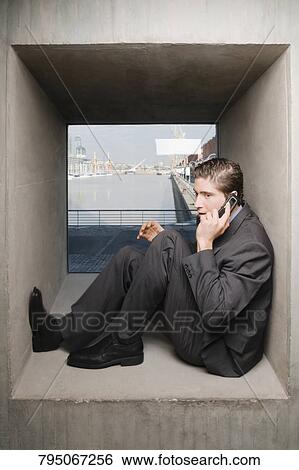 Stock Photograph - Side profile of a businessman sitting on a window sill and talking on a mobile phone. Fotosearch
