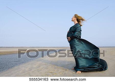 Stock Image - Side profile of a pregnant woman standing on the beach. Fotosearch