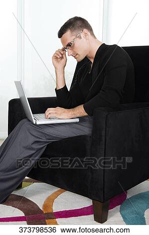 Stock Photograph - Side profile of a young man sitting in an armchair and working on a laptop. Fotosearch