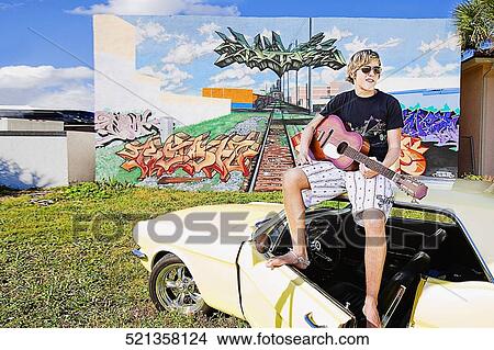 Teenage boy sitting on a car and playing guitar View Large Photo Image Picture - Teenage boy sitting on a car and playing guitar. Fotosearch