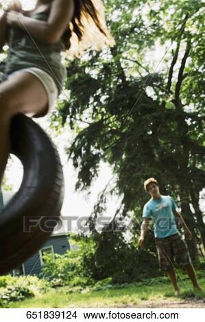 Picture - Teenage girl swinging on a tire swing with a young man in the background. Fotosearch