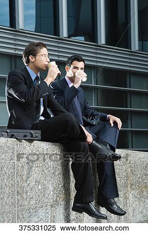 Stock Photography - Two businessmen sitting at the edge of a wall and drinking from disposable cups. Fotosearch