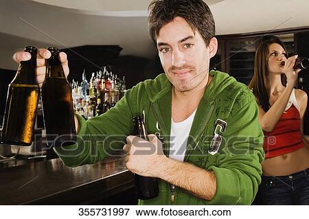 Young man holding a beer bottle at a bar counter with a young woman drinking behind him View Large Photo Image Stock Photo - Young man holding a beer bottle at a bar counter with a young woman drinking behind him. Fotosearch