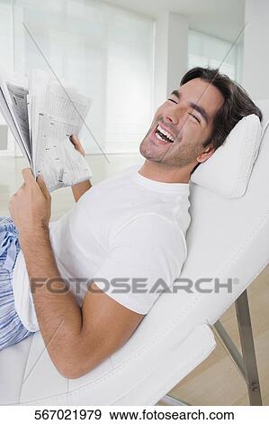 Young man holding a newspaper on a reclining chair and smiling View Large Photo Image Stock Photo - Young man holding a newspaper on a reclining chair and smiling. Fotosearch