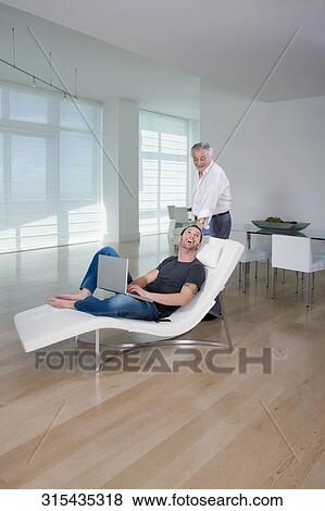 Stock Photo - Young man using a laptop with his grandfather standing behind him and smiling. Fotosearch
