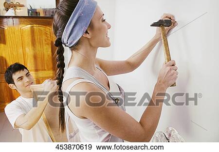 Young woman hammering nail into wall with a young man holding a picture frame View Large Photo Image Stock Photo - Young woman hammering nail into wall with a young man holding a picture frame. Fotosearch